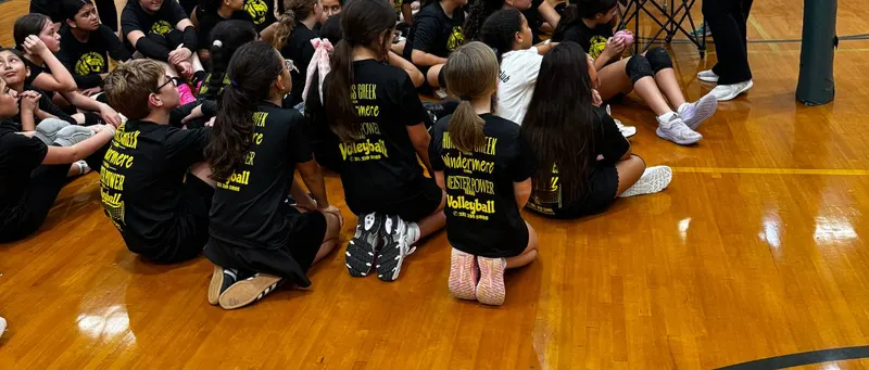 Club players sitting on a gym floor in black shirts listing Hunters Creek and Windermere
